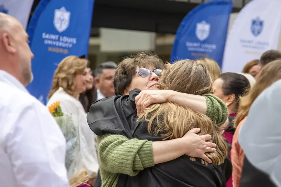 A woman hugs a participant in a graduation ceremony and looks up at the sky, while others mill around the area.